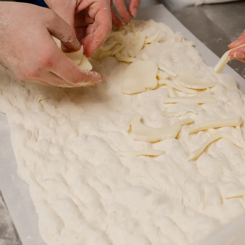 Mani che stendono e farciscono l’impasto della focaccia in teglia